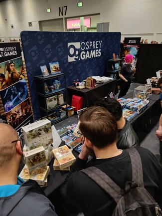 A photo of the Osprey Games stand at Salute 2023, with banners in the background and a wide selection of Osprey wargames and North Star miniatures displayed on tables and shelves.
