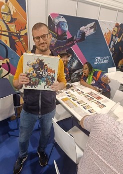 A photo of board game designer Paolo Mori at the 2024 Essen SPIEL convention, on the Osprey Games stand holding a finished copy of Battalion: War of the Ancients, with members of the BoardGameGeek team smiling in the background behind him