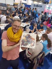A photo of Roland MacDonald at the Essen SPIEL convention 2024 on the Osprey Games stand holding a finished game box for Battalion: War of the Ancients showing his cover art depicting a diverse array of ancient soldiers and armies