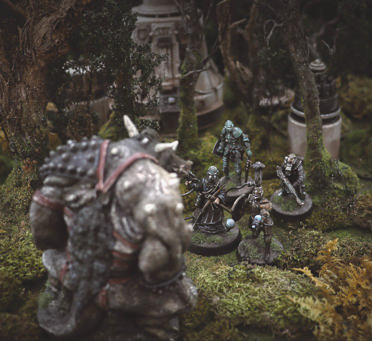 A photo of fully painted miniature figures and terrain for the apocalyptic monster hunting wargame The Doomed. In a forest growing around high-tech ruins, a horrific monstrosity towers over a band of futuristic knight-like figures.