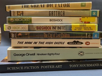 A stack of video games, DVDs and books: Science Fiction Poster Art, 1984, The Man in the High Castle, Bioshock Infinite, Bioshock, Gattaca and The Great Dictator