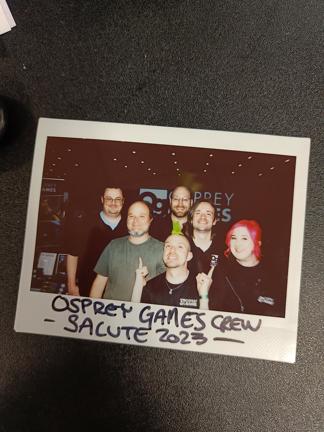 A photo of a polaroid picture of the Osprey Games team at Salute 2023 and author Joseph A. McCullough smiling in front of the Osprey Games stand.