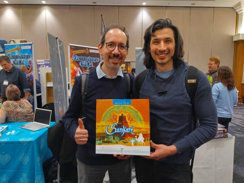A photo of designers Fabio Lopiano & Mandela Fernández-Grandon smiling holding the Sankoré box at UK Games Expo 2023