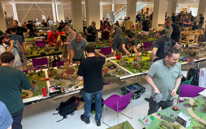A close-up photo of people smiling playing Bolt Action on tables covered with fully painted miniature terrain