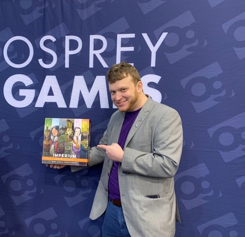 A photo of co-designer Dávid Turczi holding an early copy of Imperium: Horizons in front of an Osprey Games banner