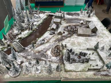 A photo of the Osprey Games demo table for The Silver Bayonet at the wargaming convention Salute 2023, with terrain and figures provided by North Star Military Figures. It is a snowy 19th-century village surrounded by forest and a river, with miniature figures of Napoleonic soldiers set up at various points.