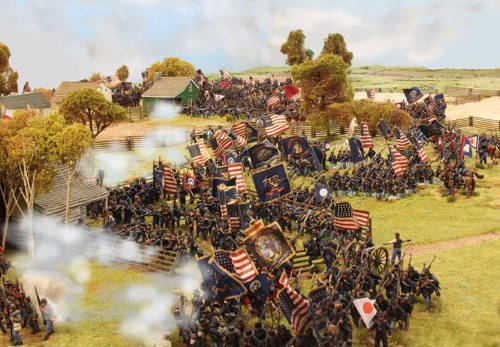 A photo of fully painted miniature figures and terrain for wargaming: a huge Union American Civil War army descending on a wooded settlement with clouds of gunsmoke hanging in the air