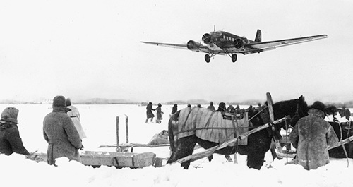 Snowy scene, horse and people in the foreground. A plane is landing in the background.