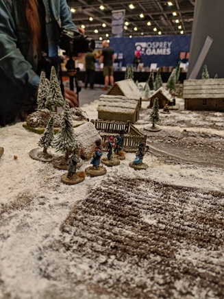 A photo of the Osprey Games The Silver Bayonet demo table at Salute 2023. It shows a small band of Napoleonic French soldiers amidst trees and a village, all covered in snow.