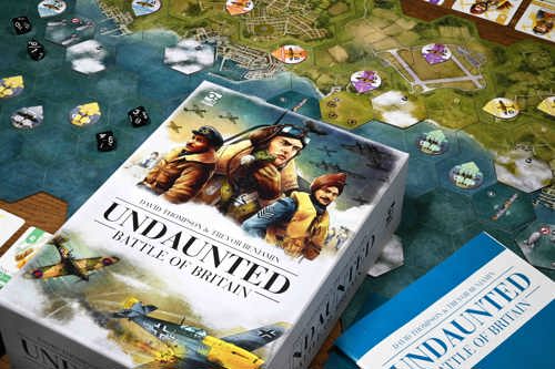 A photo of the Undaunted: Battle of Britain board game box, with cover art depicting a diverse range of WW2 pilots, laid out on top of a set-up game board with various dice, tokens and counters placed around it.