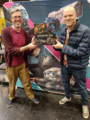 A photo of artist Roland MacDonald and co-designer Trevor Benjamin stood smiling holding a copy of Callisto in front of a themed banner at UKGE 2024
