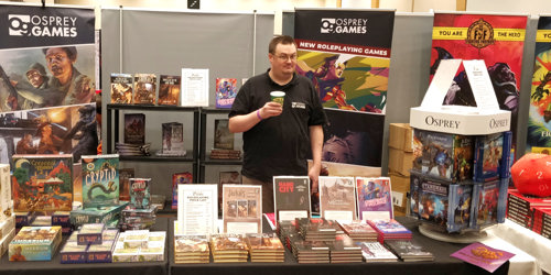 A photo of a member of the Osprey Games team stood behind our stand at Dragonmeet 2022