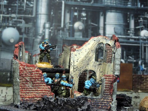 A photo of a scene laid out with miniature figures and terrain: blue-skinned alien marines are taking shelter in a bombed-out building in front of an industrial sci-fi backdrop