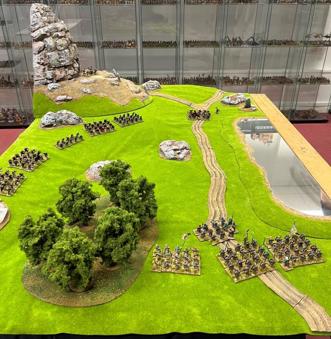 A photo of the North Star Military Figures Oathmark demo table for the wargaming convention Salute 2023. A grassy, hilly field is marked by a patch of forest, a lake, and a towering stone outcrop. Two opposing fantasy armies of miniature figures have been arrayed against each other.