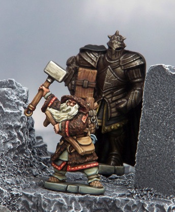 A photo of two fully painted miniature figures surrounded by snowy, mountainous terrain: an old wizard wearing furs and holding up a hammer, and a towering armoured figure stood behind him