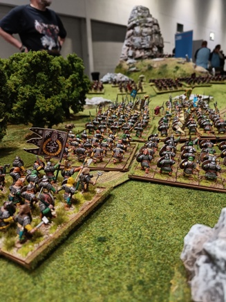 A photo of the Oathmark demo table at Salute 2023 from North Star Military Figures. It shows an army of dwarves advancing past rocks and trees against an army of goblins beneath a hill and a towering stone outcrop.