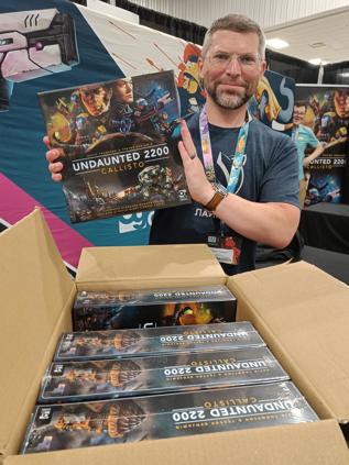 A photo of co-designer David Thompson smiling opening a box full of Callisto copies at Gen Con 2024